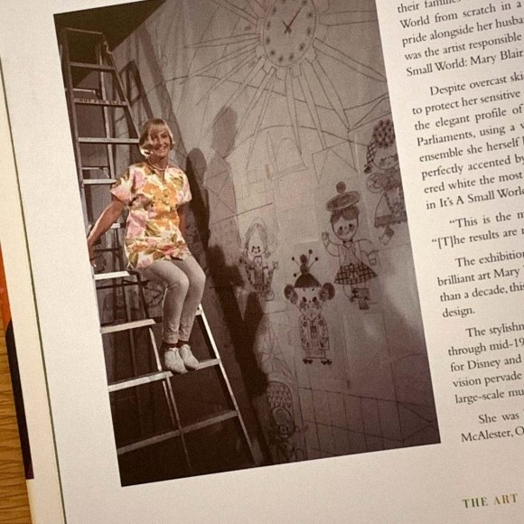 A photo of Mary Blair sitting on a ladder next to a mural and smiling at the camera. The photo is featured in 'The Art And Flair Of Mary Blair' by John Canemaker.