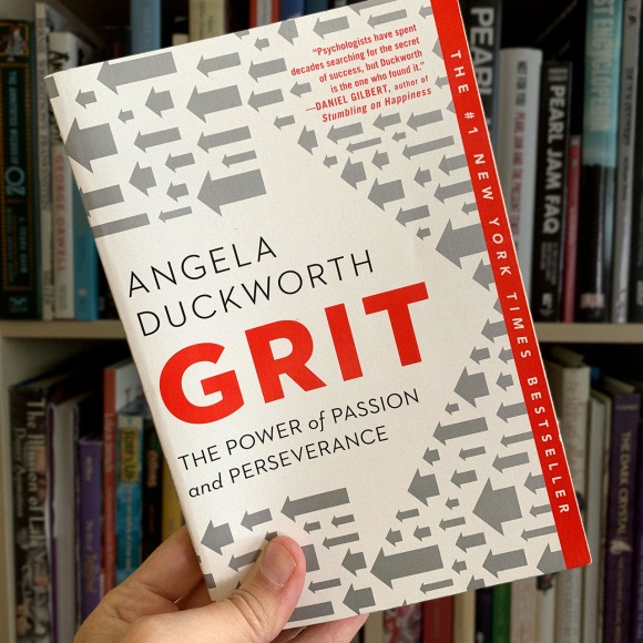 A photo of Angela Duckworth’s book ‘Grit’ being held up in front of a bookshelf.