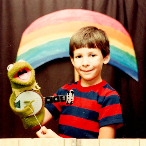 A young Chris Oatley with a vintage Kermit The Frog puppet, circa 1984. The pupped holds a handmade banjo. In the background there is a rainbow made with construction paper and painted with Crayola markers.