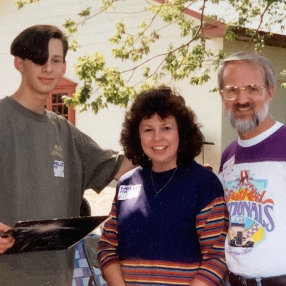 A photo of Chris Oatley as a teenager with his parents. They’re outside in the shade, smiling at the camera.