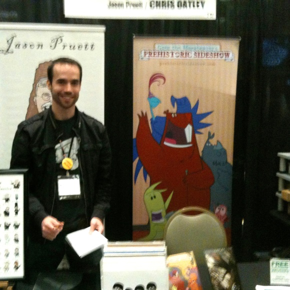 A photo of Jason Pruett and Chris Oatley’s shared booth at CTNX in 2011. Jason Pruett smiles at the camera in front of a couple of banner images, one of which is prominently featuring the dinosaur characters of Chris Oatley’s personal project, ‘Greg The Megabeaver’s Prehistoric Sideshow.’