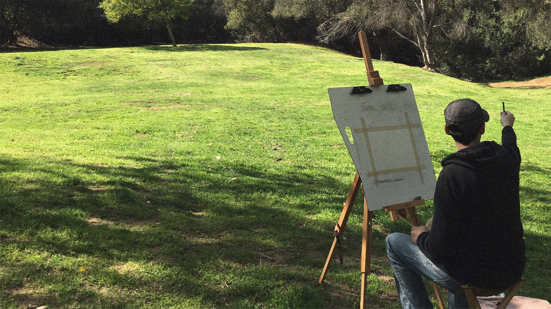 A photo of Chris Oatley sitting in the park with his easel, measuring a distant tree with his pencil for a plein air sketch.