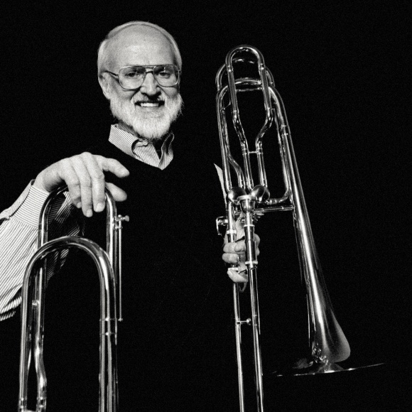 A black and white photo of Doug Oatley smiling at the camera, leaning on one trombone and holding another in his other hand.